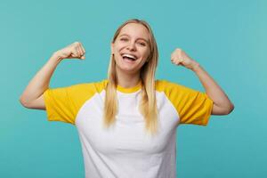 content Jeune aux yeux bleus longue aux cheveux blond femme à la recherche gaiement à caméra avec large sourire tandis que montrant fort biceps, permanent plus de bleu Contexte photo