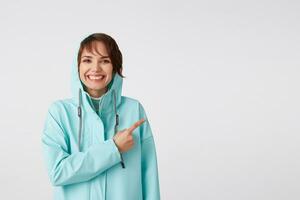 mignonne souriant cheveux courts frisé Dame dans bleu pluie manteau, veut à dessiner vous attention à le copie espace sur le droite côté et montrer du doigt à lui, des stands plus de blanc Contexte. photo