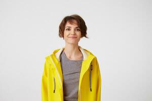 photo de positif Jeune agréable femme dans Jaune pluie manteau, prendre plaisir le vie, regards à le caméra avec content expressions, souriant plus de blanc mur.