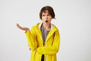 sous le choc cheveux courts Jeune femme dans Jaune pluie manteau, avec large ouvert bouche et indigné expression, en mettant le paume en haut, renfrogné et à la recherche une façon avec large ouvert bouche, des stands plus de blanc Contexte. photo