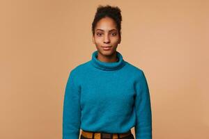 portrait de calme Jeune africain américain femme avec frisé foncé cheveux , portant une bleu chandail, isolé plus de beige Contexte. photo