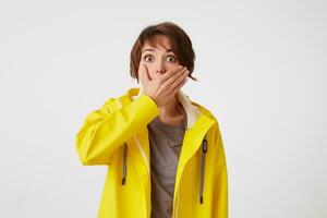 portrait de cheveux courts frisé femme dans Jaune pluie manteau, entend incroyable nouvelles , couvert bouche avec main, des stands plus de blanc mur avec large ouvert yeux avec surpris expression. photo