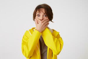 portrait de effrayé cheveux courts frisé femme dans Jaune pluie manteau, entendu une terrible histoire, couvert bouche avec palmiers, des stands plus de blanc mur avec large ouvert yeux avec peur expression. photo