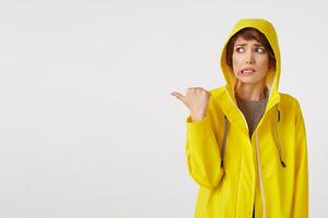 Jeune agréable femme dans une Jaune imperméable avec une dégoûté expression sur sa visage veut à dessiner votre attention à le copie espace sur le la gauche montrer du doigt avec le sien doigt, permanent plus de blanc mur. photo