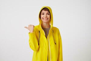 portrait de Jeune content mignonne court aux cheveux fille porte dans Jaune pluie manteau, largement sourires, veut à dessiner vous attention et points à copie espace à le gauche, des stands plus de blanc mur. photo