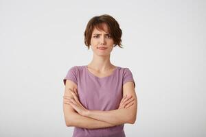 portrait de renfrogné mécontent cheveux courts Dame dans Vide T-shirt, les doutes le décision avec bras franchi, des stands plus de blanc mur. photo