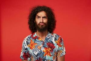 studio coup de aux yeux bruns Jeune brunette homme avec frisé cheveux grimaçant et renfrogné le sien visage contre rouge arrière-plan, portant luxuriant barbe et multicolore fleuri chemise photo