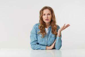 portrait de confus Jeune femme avec longue blond ondulé cheveux, séance à le tableau, un paume soulevé, regards avec scepticisme mécontent avec indignation avec incompréhension, plus de blanc Contexte. photo