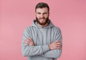 portrait de Jeune renfrogné Beau rouge barbu homme dans gris sweatshirt à capuche, des stands avec franchi bras, mal regards à caméra, dénudé le sien dents, des stands plus de rose Contexte. photo