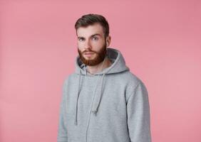 portrait de Jeune attrayant rouge barbu homme dans gris sweatshirt à capuche, regards bien et calme, sourires, écouter chanson dans écouteurs, jouit le musique, des stands plus de rose Contexte. photo