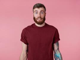 quelque chose faux Jeune attrayant tatoué rouge barbu homme dans Vide T-shirt, regards sous le choc et triste, des stands plus de rose Contexte. photo