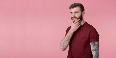 portrait de Jeune attrayant en pensant tatoué rouge barbu homme dans rouge T-shirt, regarde ailleurs et touche le menton, souriant et touche le menton, des stands plus de rose Contexte avec espace de copie. photo