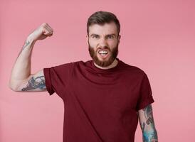 Jeune mal rouge barbu homme dans Vide T-shirt, des stands plus de rose arrière-plan, regards cool, prendre plaisir lui-même et démontrer puissant, regards à le caméra et en hurlant. photo