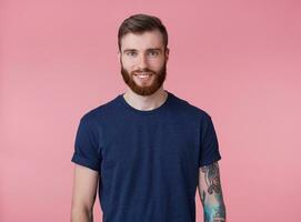 portrait de Jeune content attrayant à barbe rousse gars avec bleu yeux, portant une bleu T-shirt, souriant et à la recherche à le caméra isolé plus de rose Contexte. photo