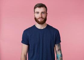 Jeune attrayant à barbe rousse gars avec bleu yeux, portant une bleu T-shirt, à la recherche à le caméra avec une calme faciale expression isolé plus de rose Contexte. photo