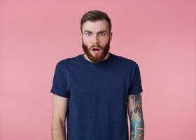 photo de Jeune Beau étonné rouge barbu hommehomme dans Vide T-shirt, entend incroyable nouvelles, regards surpris, des stands plus de rose Contexte avec large ouvert bouche et yeux.