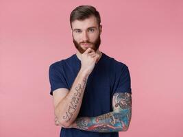 pensif attrayant à barbe rousse gars avec bleu yeux, portant une bleu T-shirt, détient le sien main à le sien menton et regards pensivement dans le caméra isolé plus de rose Contexte. photo