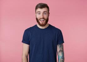 Jeune content attrayant à barbe rousse Jeune gars, portant une bleu T-shirt, à la recherche à le caméra avec large ouvert bouche et yeux dans surprendre, vu quelque chose mignon, isolé plus de rose Contexte. photo