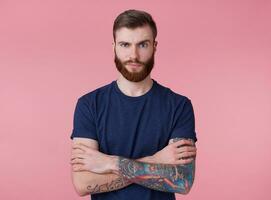 portrait de mécontent Jeune à barbe rousse Beau courageux gars avec franchi bras, regards indigné, à la recherche à le caméra avec élevé sourcil, isolé plus de rose Contexte. photo