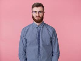 triste Jeune Beau rouge barbu homme avec des lunettes et une rayé chemise, triste cette le fin de le travail la semaine est ne pas bientôt, et il est déjà très fatigué. des stands plus de rose Contexte. photo