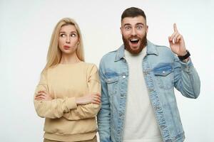 étonné Jeune Beau barbu brunette gars élevage indice doigt et à la recherche hébété à caméra avec large yeux et bouche ouvert, posant plus de blanc Contexte avec perplexe jolie blond Dame photo