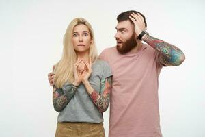 perplexe Jeune jolie blond tatoué Dame mordant inquiétant lèvre inférieure tandis que à la recherche confusément à caméra, posant plus de blanc Contexte avec perplexe brunette barbu Masculin photo