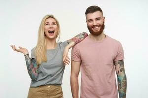 horizontal coup de Jeune attrayant tatoué couple étant dans haute esprit et souriant largement tandis que posant plus de blanc Contexte dans gris et rose de base t-shirts photo