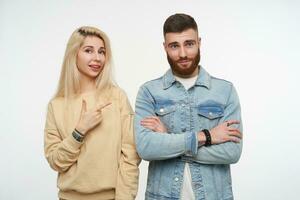 studio photo de Jeune jolie blond femelle dans beige sweat-shirt montrer du doigt positivement sur mignonne Jeune marron aux cheveux barbu homme avec plié mains, posant plus de blanc Contexte