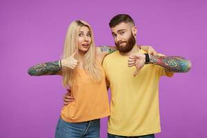 studio photo de Jeune aux yeux bleus tatoué jolie couple montrant comme et Ne pas aimer tandis que à la recherche à caméra et étreindre chaque autre, isolé plus de violet Contexte