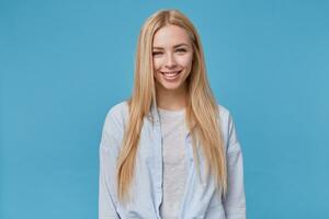 portrait de Jeune blond femelle avec décontractée coiffure posant plus de bleu arrière-plan, portant bleu chemise et gris T-shirt, montrant positif émotions après recevoir agréable compliment photo