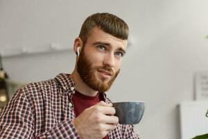 photo de Jeune attrayant gingembre barbu homme en portant une gris café tasse, rêveusement regards en haut et jouit café, portant dans de base vêtements.