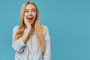 content Jeune blond femme avec longue cheveux portant décontractée vêtements, montrant positif émotions après recevoir agréable compliment, posant plus de bleu Contexte et en gardant paume sur joue photo