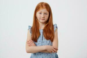 portrait de peu malheureux taches de rousseur roux fille avec deux queues, mécontent regards à le caméra, porte dans bleu robe, des stands avec franchi bras plus de blanc Contexte. photo