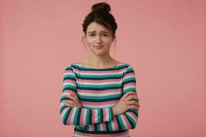 adolescent fille, déçu femme avec brunette cheveux et chignon. portant rayé chemisier et plis sa mains sur une poitrine. émotif concept. en train de regarder à le caméra isolé plus de pastel rose Contexte photo