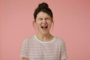 adolescent fille, en colère à la recherche femme avec brunette cheveux et chignon. portant T-shirt avec rouge bandes et en hurlant dans colère. émotif concept. supporter isolé plus de pastel rose Contexte photo