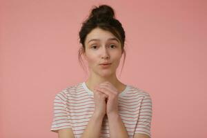 portrait de attractif, brunette fille avec brunette cheveux et chignon. portant T-shirt avec rouge bandes et mendicité toi. émotif concept. en train de regarder à le caméra isolé plus de pastel rose Contexte photo