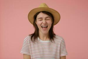 adolescent fille, content à la recherche femme avec longue brunette cheveux. portant T-shirt avec rouge bandes et chapeau. émotif concept. cris avec fermé yeux. supporter isolé plus de pastel rose Contexte photo