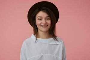 adolescent fille, joyeux, satisfait à la recherche femme avec longue brunette cheveux. portant blanc chemisier et noir chapeau. émotif concept. en train de regarder à le caméra isolé plus de pastel rose Contexte photo