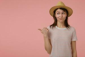 agréable à la recherche femme, magnifique fille avec longue brunette cheveux. portant T-shirt avec rouge bandes et chapeau. émotif concept. en train de regarder et montrer du doigt à le la gauche à copie espace plus de pastel rose Contexte photo