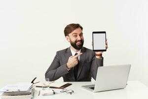 positif Jeune barbu brunette homme avec court la Coupe de cheveux séance à table plus de blanc Contexte avec tablette PC dans main, portant gris costume et à la recherche à caméra avec large sourire photo