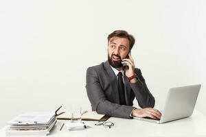 perplexe Jeune brunette homme avec barbe à la recherche de côté avec confus visage et froissement le sien front, fabrication appel avec téléphone intelligent tandis que séance à table avec mains sur clavier de le sien portable photo