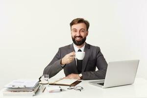 intérieur photo de positif Jeune barbu brunette homme ayant bien travail journée et souriant Heureusement à caméra tandis que séance à table plus de blanc arrière-plan, en portant tasse de café pendant Pause