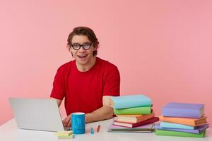 photo de content Jeune étudiant dans des lunettes porte dans rouge T-shirt, homme est assis par le table et travail avec portable et livres, largement sourires, regards content, isolé plus de rose Contexte.