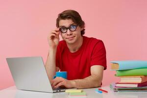 Jeune étudiant dans des lunettes porte dans rouge T-shirt, homme est assis par le table et travail avec ordinateur portable, regards à le caméra par des lunettes avec confus expression, isolé plus de rose Contexte. photo
