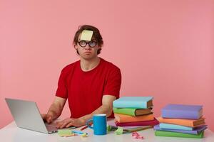 Jeune en pensant homme dans des lunettes porte dans rouge T-shirt, avec une autocollant sur le sien front, est assis par le table et travail avec carnet et livres, regards en haut et supposer, isolé plus de rose Contexte. photo