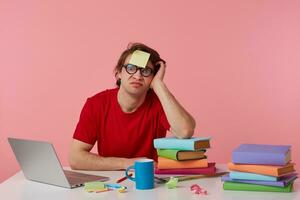 Jeune homme dans des lunettes porte dans rouge T-shirt, avec une autocollant sur le sien front, homme est assis par le table et travail avec portable et livres, isolé plus de rose Contexte. regards mécontent et malheureux. photo