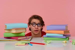 triste Jeune homme dans des lunettes porte dans rouge T-shirt, cache à le table avec livres, isolé plus de rose Contexte. regards mécontent et malheureux. photo