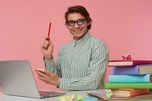 Jeune de bonne humeur gars dans des lunettes est assis par le table et travail avec ordinateur portable, détient dans main une crayon, avoir une cool idée, regards à le caméra et largement souriant, isolé plus de rose Contexte. photo