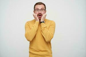 effrayé Jeune brunette barbu gars dans lunettes à la recherche à caméra avec large yeux et bouche ouvert, en gardant mains sur le sien visage tandis que permanent plus de blanc Contexte photo