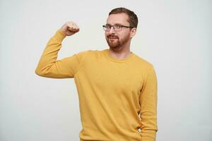 portrait de de bonne humeur Jeune barbu Masculin avec marron court cheveux à la recherche à caméra en toute confiance tandis que démontrant Puissance dans élevé main, permanent contre blanc Contexte dans décontractée vêtements photo